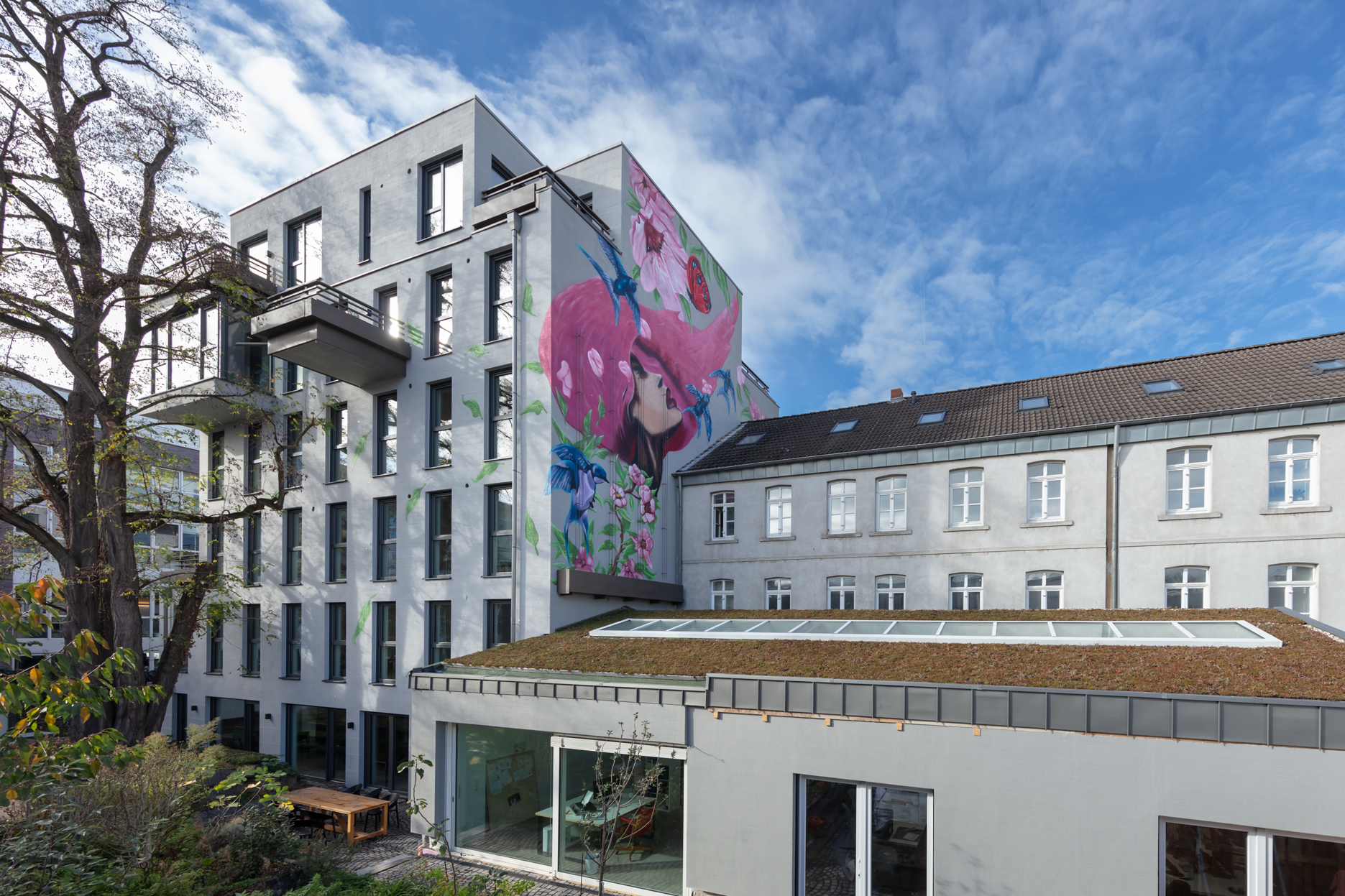 Blick auf einen Gebäudekomplex aus einem mehrstöckigen Neubau links, damit verbunden rechts ein Altbau. Im Vordergrund ein begrünter Hof mit Baum.